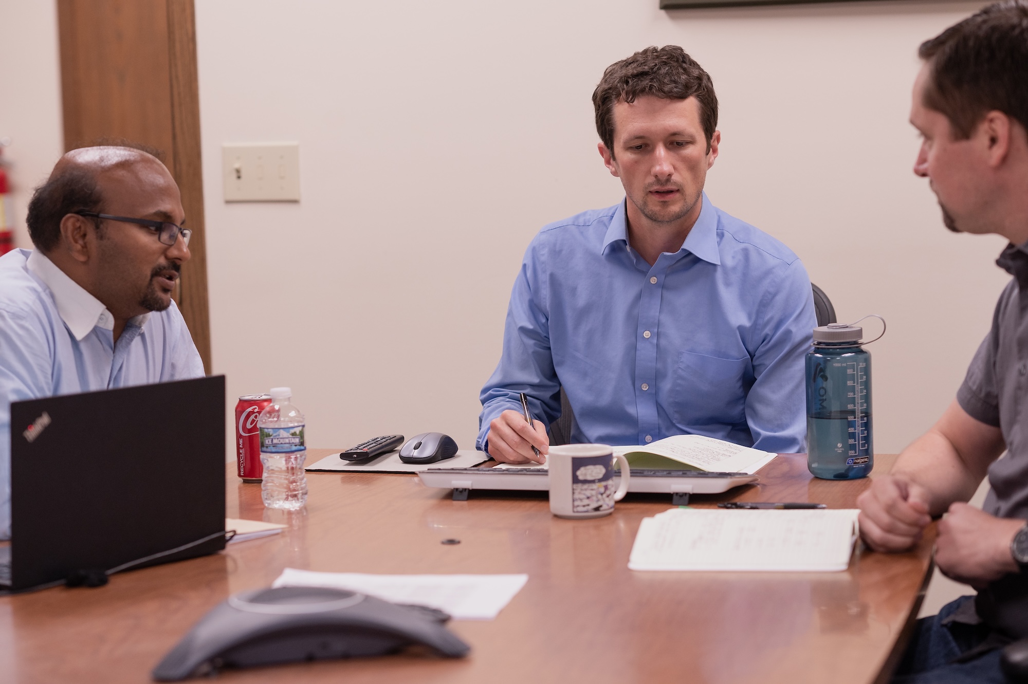 Three colleagues collaborating around a conference table, with one taking notes in an open notebook while the others engage in discussion, with a laptop, documents, and drinks on the table.